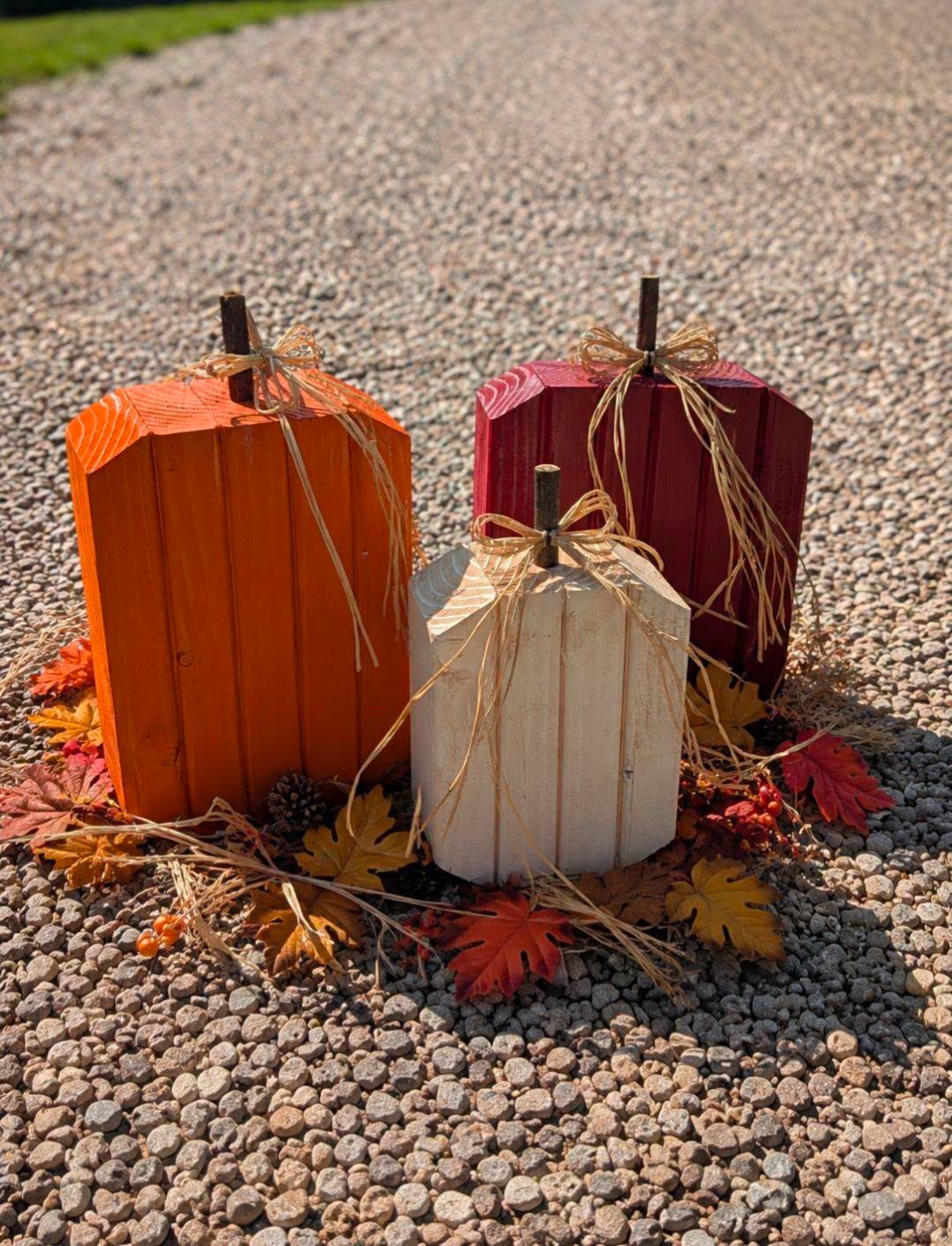 Chunky handcrafted wood pumpkins in orange, cream, and wine with branch stems and raffia bows, arranged with fall leaves